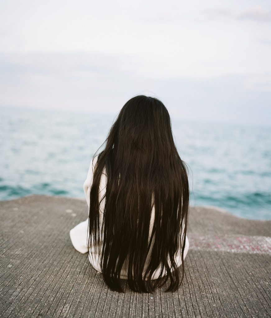 Image: Jin Lee, Untitled, 2018. A young girl sitting at the edge of Lake Michigan, seen from the back. Her long dark hair flows down to the concrete ground. Photo courtesy of the artist.
