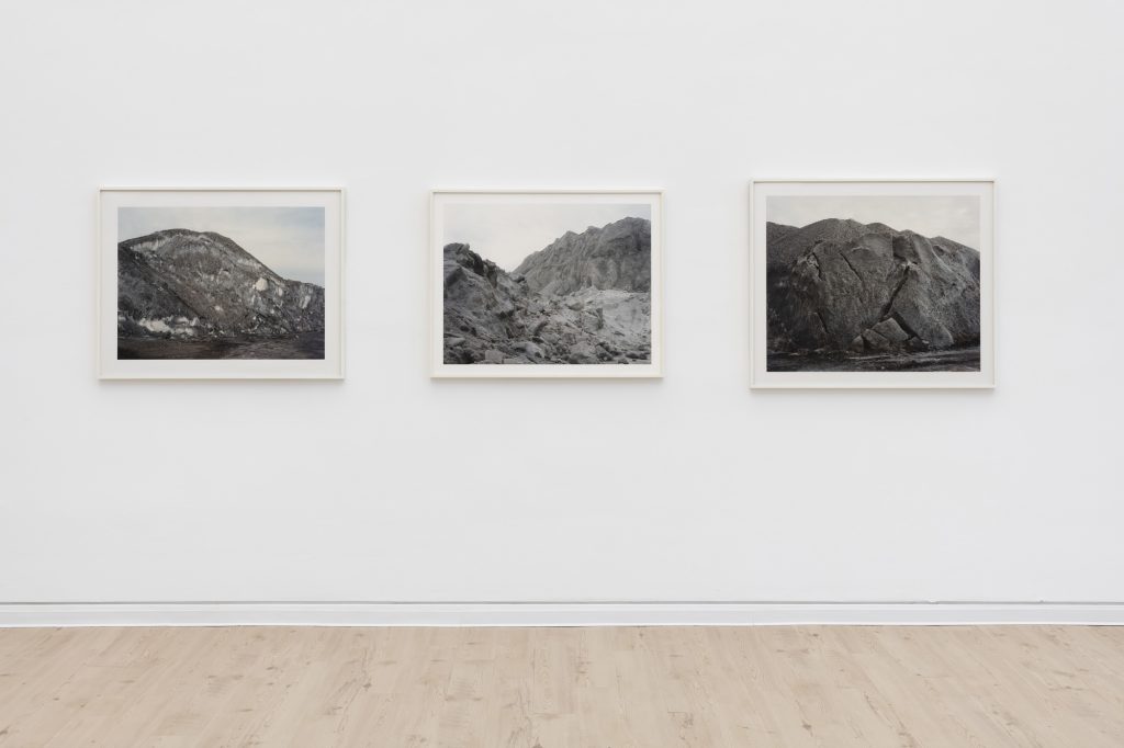 Image: Installation view of Jin Lee's series 'Salt Mountain'. Three framed photographs of gray salt mountains, seen from different angles, mounted on a white wall. Photo by James Prinz, courtesy of the Chicago Cultural Center.