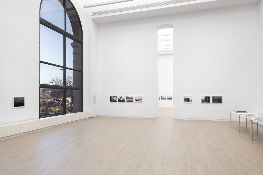 Image: Installation view of Jin Lee's exhibit 'Views & Scenes'. A large gallery with a tall, narrow doorway leading to the next gallery holds photographs from the 'Train Views' series. There is a large arched window to the east and a long wooden table with a red portfolio and prints to the west. A selection of images from 'Train Views' are mounted on the lower half of the gallery’s white walls. Photo by James Prinz, courtesy of the Chicago Cultural Center.