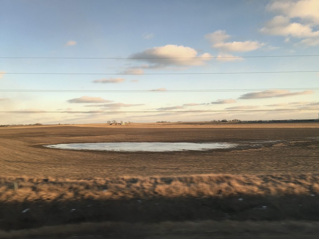Image: Jin Lee, Train View (Pond 1), 2018. A photo of a Midwestern farm at sunset. The grass is golden brown and in the center of the field, a pool of rainwater creates a transient pond. Photo courtesy of the artist.