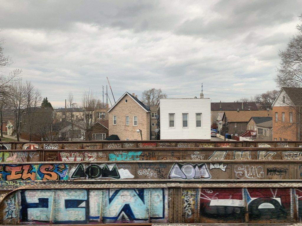 Image: Jin Lee, Train View (Two Houses), 2019. A photo of a row of houses built by the train tracks, set against a cloudy gray sky. They face front, sit side by side and differ in architectural styles. In the lower quadrant, a graffiti tagged overhead train track cuts across the image horizontally. Photo courtesy of the artist.