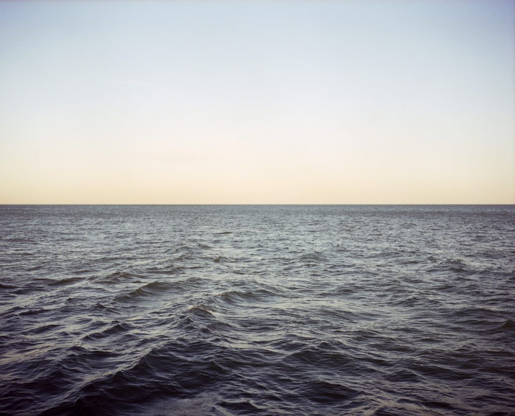 Image: Jin Lee, Great Water 8, 2017. A photograph of Lake Michigan at dusk. The frame is divided into two equal horizontal parts, with water below and sky above. The lake is choppy and dark blue, and at the horizon there is a faint orange glow. Photo courtesy of the artist.