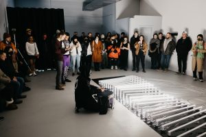 Featured image: Amay Kataria, Supersynthesis, 2022, Mu Gallery Chicago. Artist Sal Moreno performs in the middle of a crowd. Sal Moreno is kneeling on the floor playing a snare drum in front of an installation of horizontal white bars. Image courtesy of the artist.
