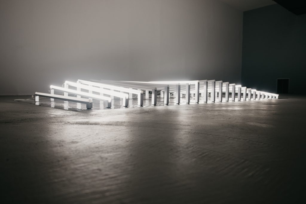 Image: Amay Kataria, Supersynthesis, 2022, MuGallery Chicago. Installation view of Supersynthesis: more than 20 LED light tubes are laid out on the floor like bed slats. Each light is raised slightly above the floor at subtly different height and angularity. Together they form a curved light plane. Image courtesy of the artist.