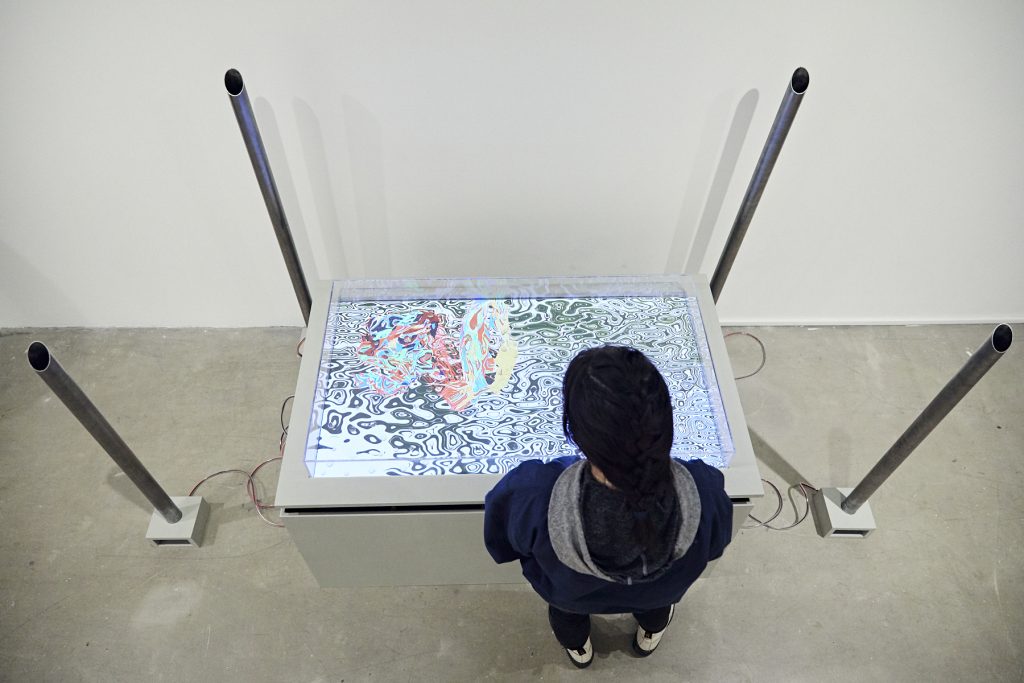Image: Amay Kataria, Figments of Desire, 2019. A bird's-eye view of the work with an audience member standing next to the installation. A big screen that displays generative patterns is laid flat on top of a waist-high pedestal. Four metal tubes are each perpendicularly pinned to a device on the ground to help amplify the sound component of the work. Image courtesy of the artist.