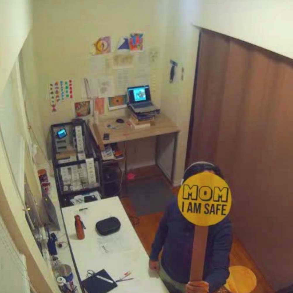 Image: Amay Kataria, Momimsafe, 2020, online performance documentation. The artist himself is seen in his studio caught by what appears to be a fish-eyed camera installed high on the wall. Where the artist's face is in the image is covered by a yellow round sticker that reads "MOM I AM SAFE." Image courtesy of the artist.