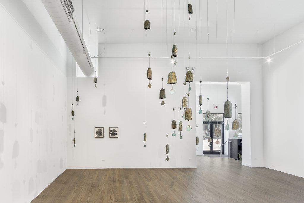 Image: Daniel G. Baird, KNELL series, 2021, and Kaveri Raina, Untitled (To Hover), 2020. A brightly lit gallery interior with white walls, this is the second room of PATRON’s gallery space. A series of brass bells hanging at various heights occupy the central space of the gallery. Their clappers have drop shapes extending down from the bell that catch shadows and reflections from ambient light. On the far left corner, the shadows of the bells guide the eye to two small, framed graphite works on paper. Image courtesy the artist and PATRON Gallery, Chicago. Photography by Evan Jenkins.