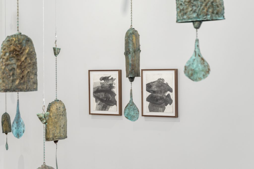 Image: (at center) Kaveri Raina, Untitled (To Hover), 2020. Two graphite on paper drawings of ambiguous shapes and figures moving in all directions, yet weighted in the center of the composition, sit side by side in walnut frames. In the foreground, out of focus details of oxidized brass and bronze from Baird’s KNELLs fill the gaps. Image courtesy the artist and PATRON Gallery, Chicago. Photography by Evan Jenkins.