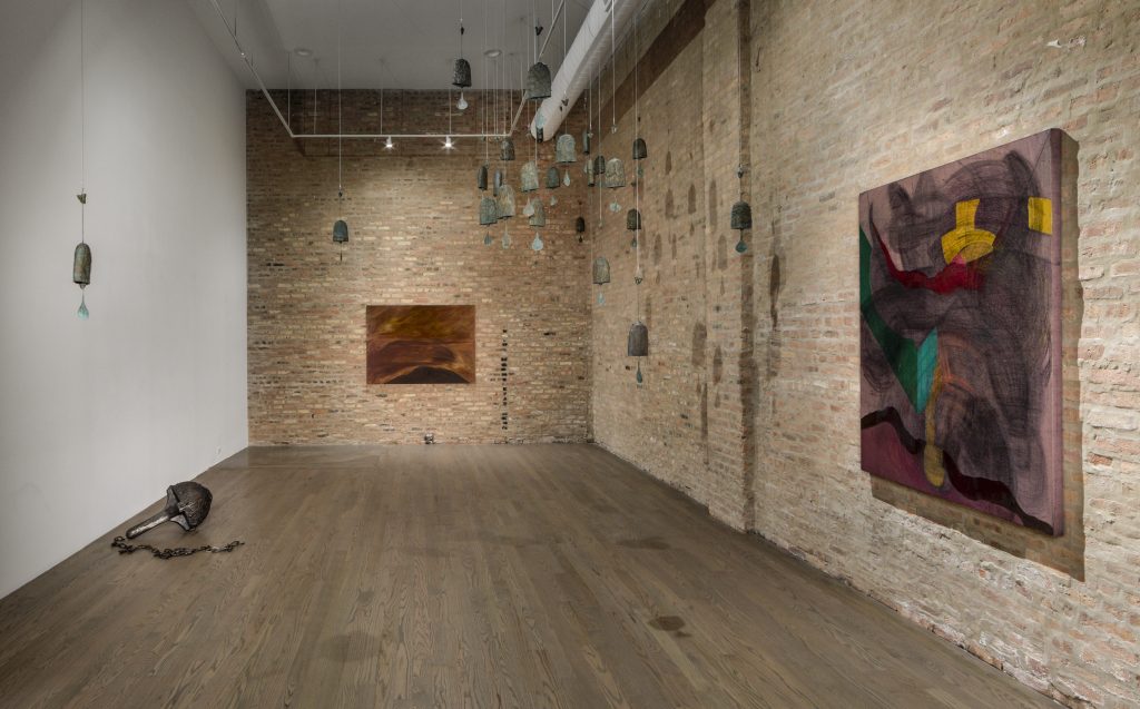 Image: Installation view of the final gallery room of Heft. An atmospheric, dimly lit gallery with gray oak wood floors has one white wall to the left hand side, and two exposed red brick walls at the far and right wall. Knowles' piece The Solemn and Dignified Burial Befitting My Beloved for All Seasons, 2022, hangs spotlit at the far wall. A broken, black stain runs down the wall, a foot to the right of the painting. On the right, we see Raina’s Revolt; Internal Brewing Slow Rage, 2021, and in between the two, a series of Baird’s KNELL bells hang down from the ceiling at undulating, steeper heights. To the left is an iron and steel ball held by a claw and chain titled, Anchor, 2022, by Baird. Image courtesy the artist and PATRON Gallery, Chicago. Photography by Evan Jenkins.