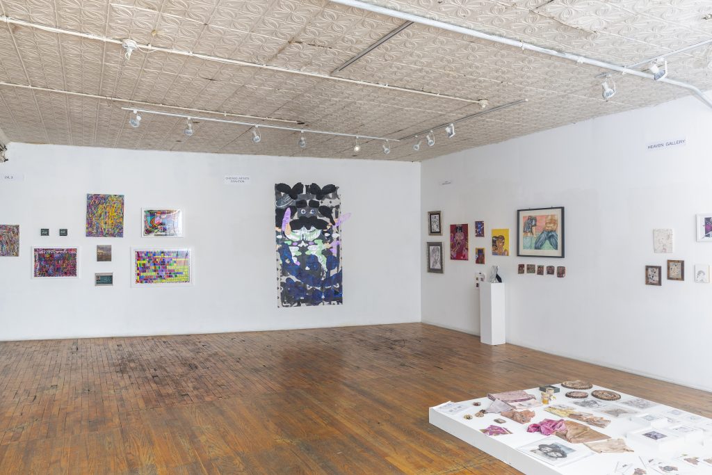 Image: Installation view of Nubes Art Fair at Heaven Gallery, 2022. Multi-color paintings and drawings hang on the walls; a sculpture sits on a plinth on the right wall and a white platform, estimated less than a one-foot tall lays on the floor with drawings and sculptures laid flat on top.