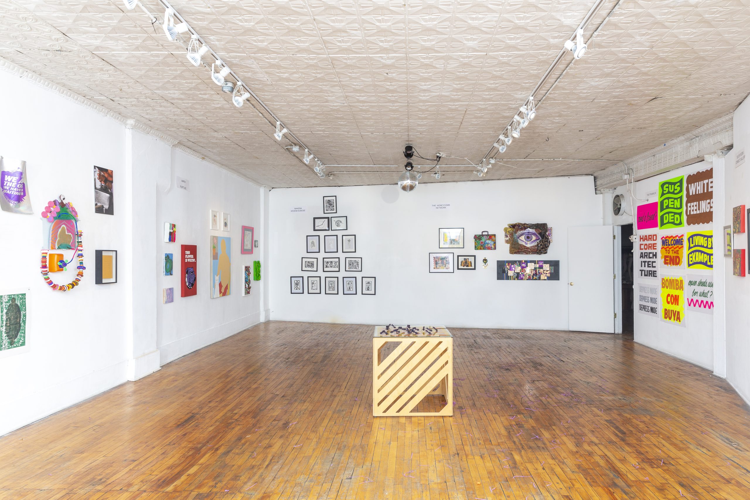 Image: Installation view of Nubes Art Fair. A golden-yellow cube sculpture with diagonable stripes sits in the center of the gallery room. Paintings, drawing and multi-media works hang on the wall.