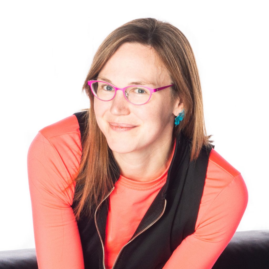 Image: A photo of Marya Spont-Lemus, shown from the chest up and smiling slightly. Spont-Lemus, a white person, wears a black top open over a salmon pink long-sleeved shirt, as well as pink glasses and teal earrings. She appears against a black and white background. Photo by Lucas Anti.