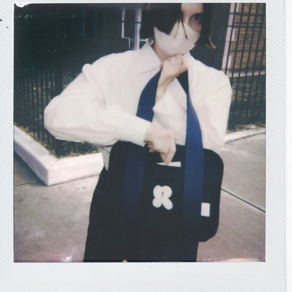 Image: A polaroid photo of a person outside a black iron gate. They wear a white mask and a white shirt, their dark hair parted. They're holding a bag and looking at the camera. Photograph by Olivia Rehm.