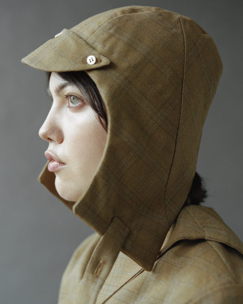 Image: On a grey background, a model wears a brown-green checked cap with flaps running down the side of their head, just past their chin, and a bill that extends just past their forehead. They wear a beige jacket. Model Ona Sian. Photograph by Brody Boggs.