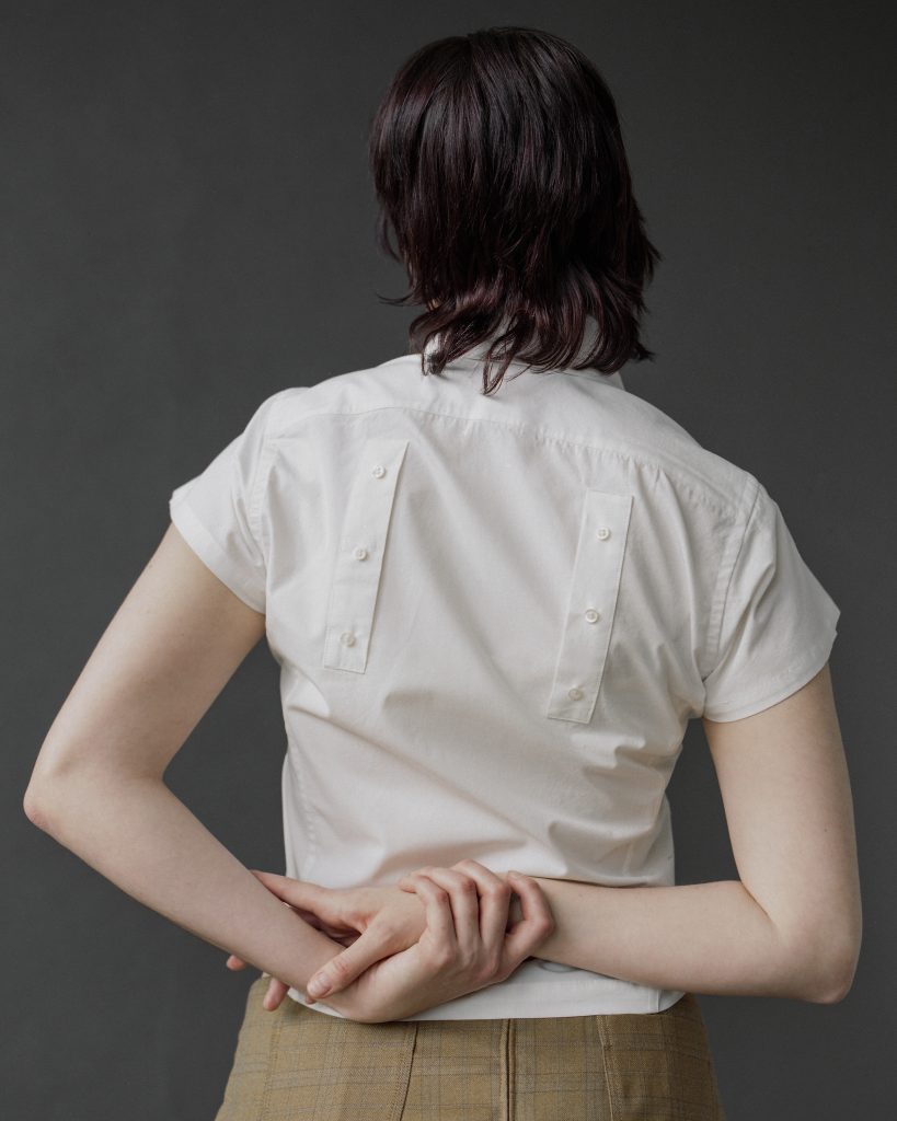 Image: On a grey background, a model wearing a white shirt holds their wrists behind their back. They wear a white shirt with two vertical strips of cloth, each with three small, white buttons running the length of them. Model Ona Sian. Photograph by Brody Boggs.