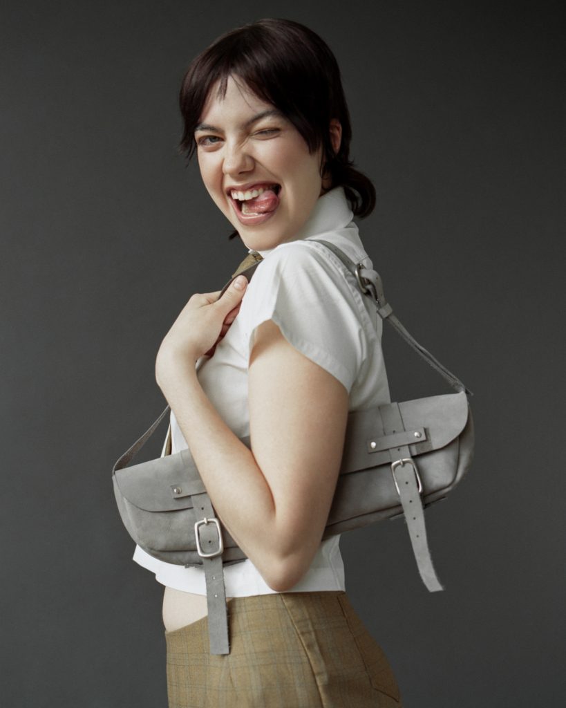 Image: On a grey background the model sticks their tongue out and closes one eye. They wear a white shirt and plaid brown legwear. Under their left arm is a long, grey purse with silver buckles. Model Ona Sian. Photograph by Brody Boggs.
