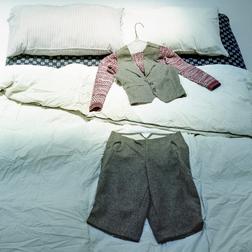 Image: A bed with white pillows at the head and white sheets. Separated by a gentle fold in the sheets, is a grey vest with magenta and white sleeves with a hanger in its neck on the upper half of the bed and, on the lower half in shade, grey short pants. Photo by Brody Boggs.