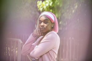 Featured image: Zarria Alexander stands with her body facing the left side of the image. She's wearing a pink jacket and a white headband. Her hair is in a pink, short bob. She has her right arm raised with her hand resting under her chin. The background of the image is abstracted with hues of pink. Photo by Kristie Kahns