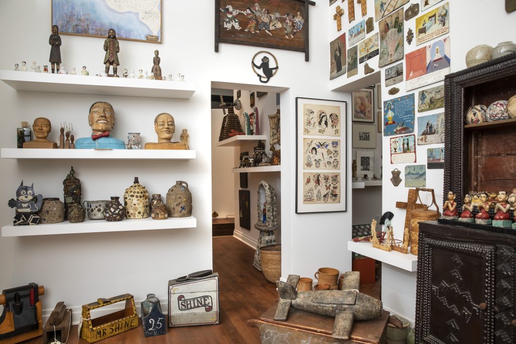 Image: Installation view of works from the Ray Yoshida Collection at the Art Preserve, 2021. Two rectangular passage ways at the center of the photo. Surrounding them, on the ground, the walls, and shelving are a variety of objects, such as ceramic pots, wooden busts, figurines, toolboxes, postcard prints, and much much more. Photo: Rich Maciejewski, courtesy of John Michael Kohler Arts Center.