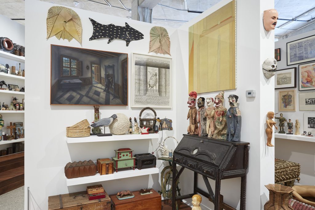 Image: Installation view of works from the Ray Yoshida Collection at the Art Preserve, 2021. A corner of the exhibit. There are Punch & Judy style puppets atop a black wooden bureau, among many many other objects. Photo: Rich Maciejewski, courtesy of John Michael Kohler Arts Center.