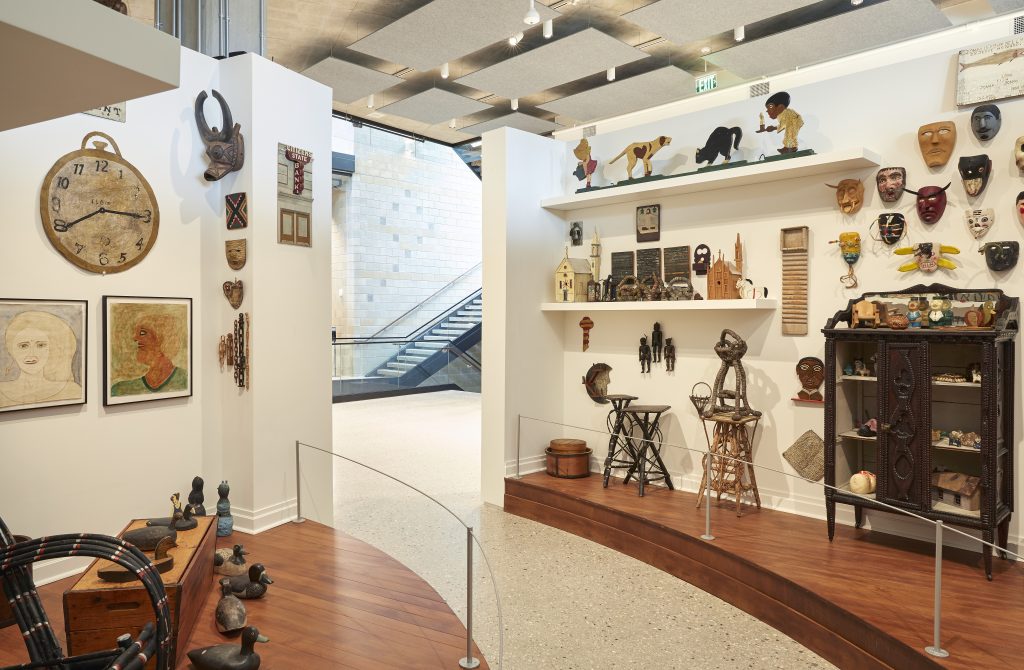 Image: Installation view of works from the Ray Yoshida Collection at the Art Preserve, 2021. A walkway through two wooden floor display sections. Each side is populated by a variety of masks, cabinets, chairs, and wooden figurines. Photo: Rich Maciejewski, courtesy of John Michael Kohler Arts Center.