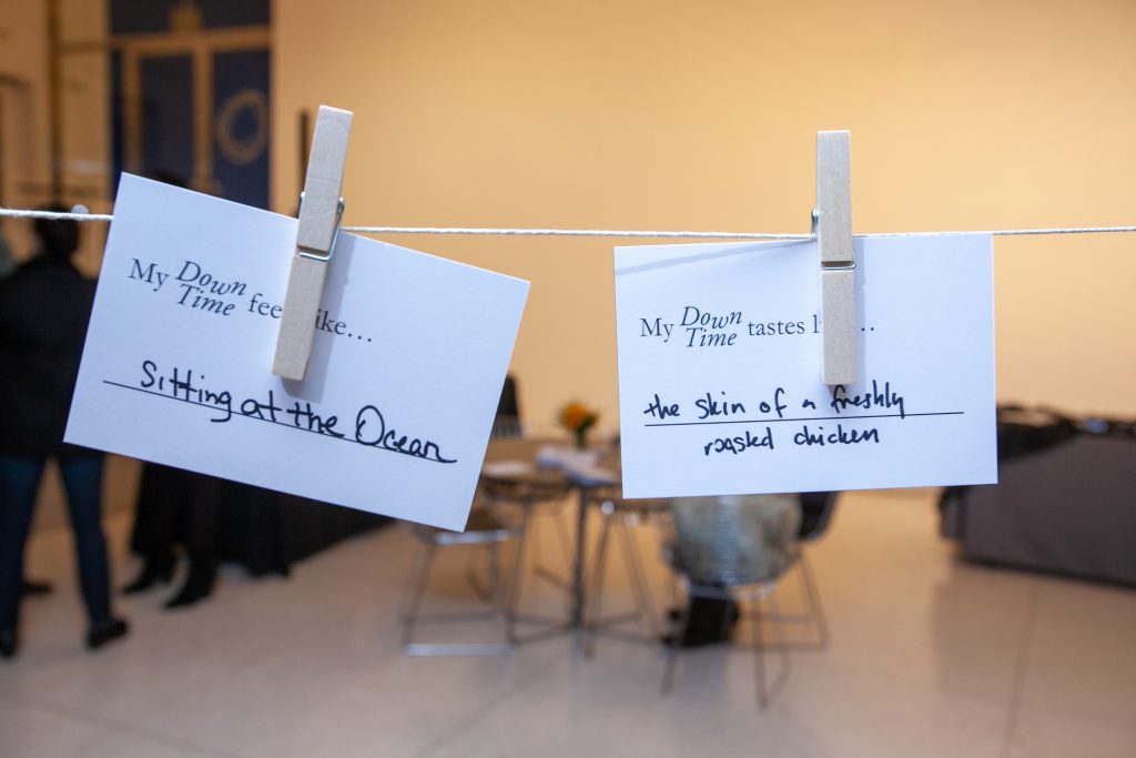 Image: Two cards are pinned with clothespins on a clothesline. The first card reads "My Down Time feels like...sitting at the ocean" the second card reads "My Down Time tastes like...the skin of a freshly roasted chicken" Opening reception of the student-curated exhibition "Down Time: On the Art of Retreat," at the Smart Museum of Art at the University of Chicago on Thursday, October 24, 2019. Photography by Eddie Quinones.