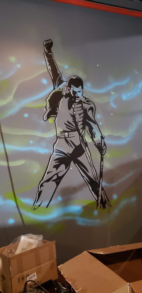 Image: A painting by Matt Sopron of music icon Freddie Mercury, seen on a wall inside of a concert venue. The image is in black outline, and is based on a famous photograph of Mercury performing at Wembley Stadium in London in 1986. The musician has his right arm raised with a clenched fist, his gaze looking down, while he clasps a microphone stand in his left hand. Photo courtesy of Matt Sopron.