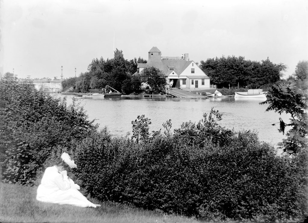 Image: A black and white photo of Jackson Park Harbor, ca: 1900. A woman in a dress sits on the grass on the left. A lake is in the background with a boathouse and several boats. Courtesy of Washington Park Camera Club.