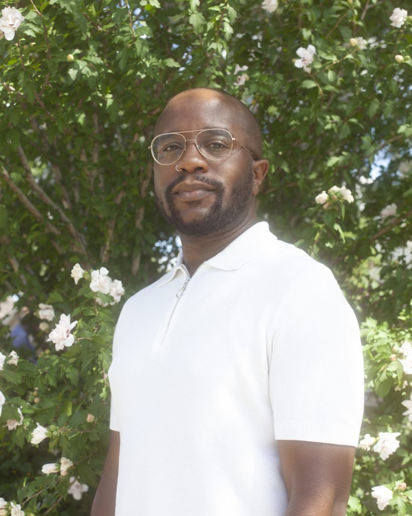 Image: Steven D. Booth stands in front of a tree that is sprinkled with white flowers, he's looking directly into the camera. Photo by Samantha Friend Cabrera.