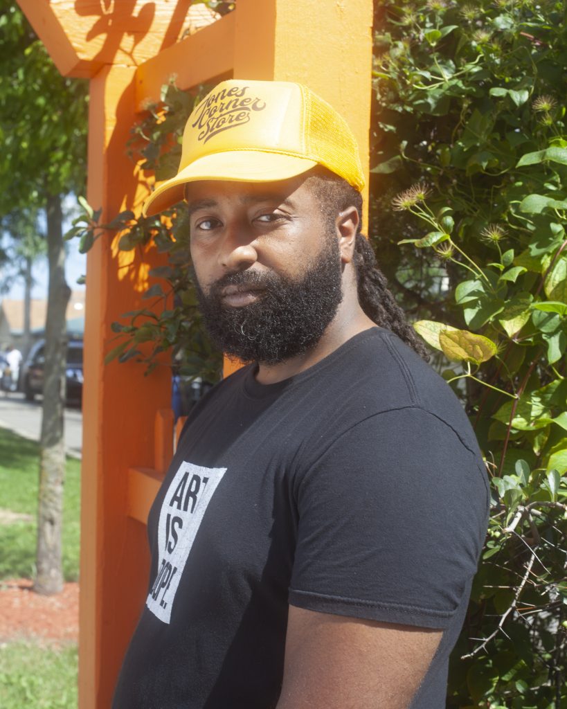 Image: Jay Simon stands in front of an orange structure and the bright green leaves of a flower bush. He wears a bright orange hat and black t-shirt that says "Art is Dope." Photo by Samantha Friend Cabrera.