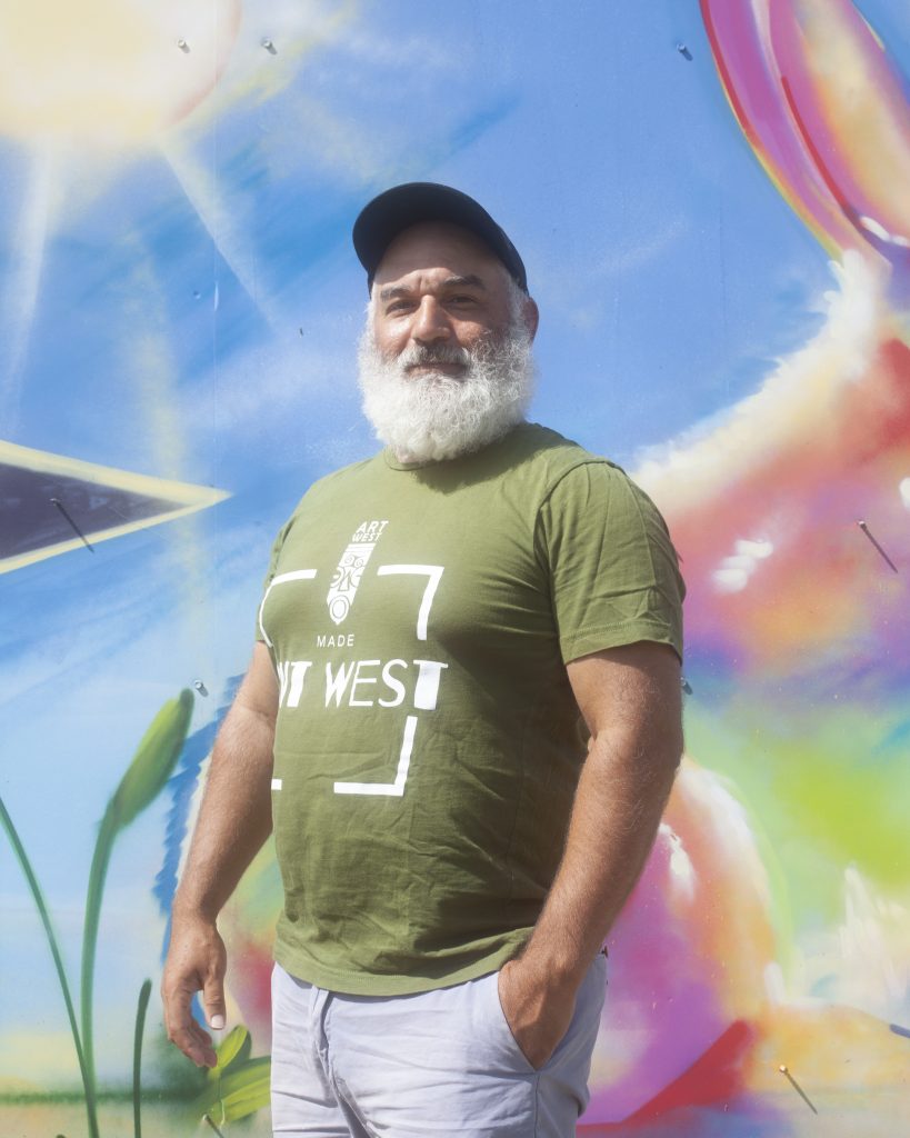 Image: Jonathan Kelley stands in front of a colorful mural, looking directly into the camera. You can see the words" Art West" on his t-shirt. Photo by Samantha Friend Cabrera.