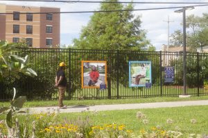 Image: Jay Simon stands on the sidewalk looking at two framed portraits he made of North Lawndale residents, the photos hanging on a fence outside. A building can be seen in the distance with grass and flowers in the foreground. Photo by Samantha Friend Cabrera.