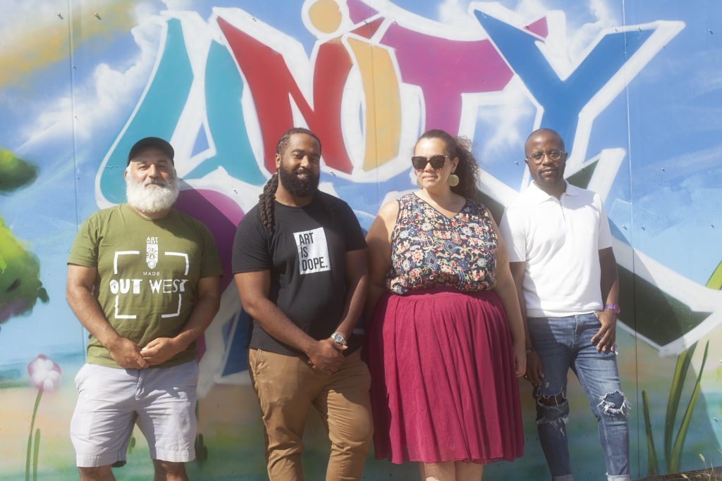 Image: Jonathan Kelley, Jay Simon, Chelsea Ridley, and Steven D. Booth stand side-by-side in front of a colorful mural with the word "Unity" painted just above their heads. They're each looking directly into the camera as the sun continues to shines bright and high over them. Photo by Samantha Friend Cabrera.