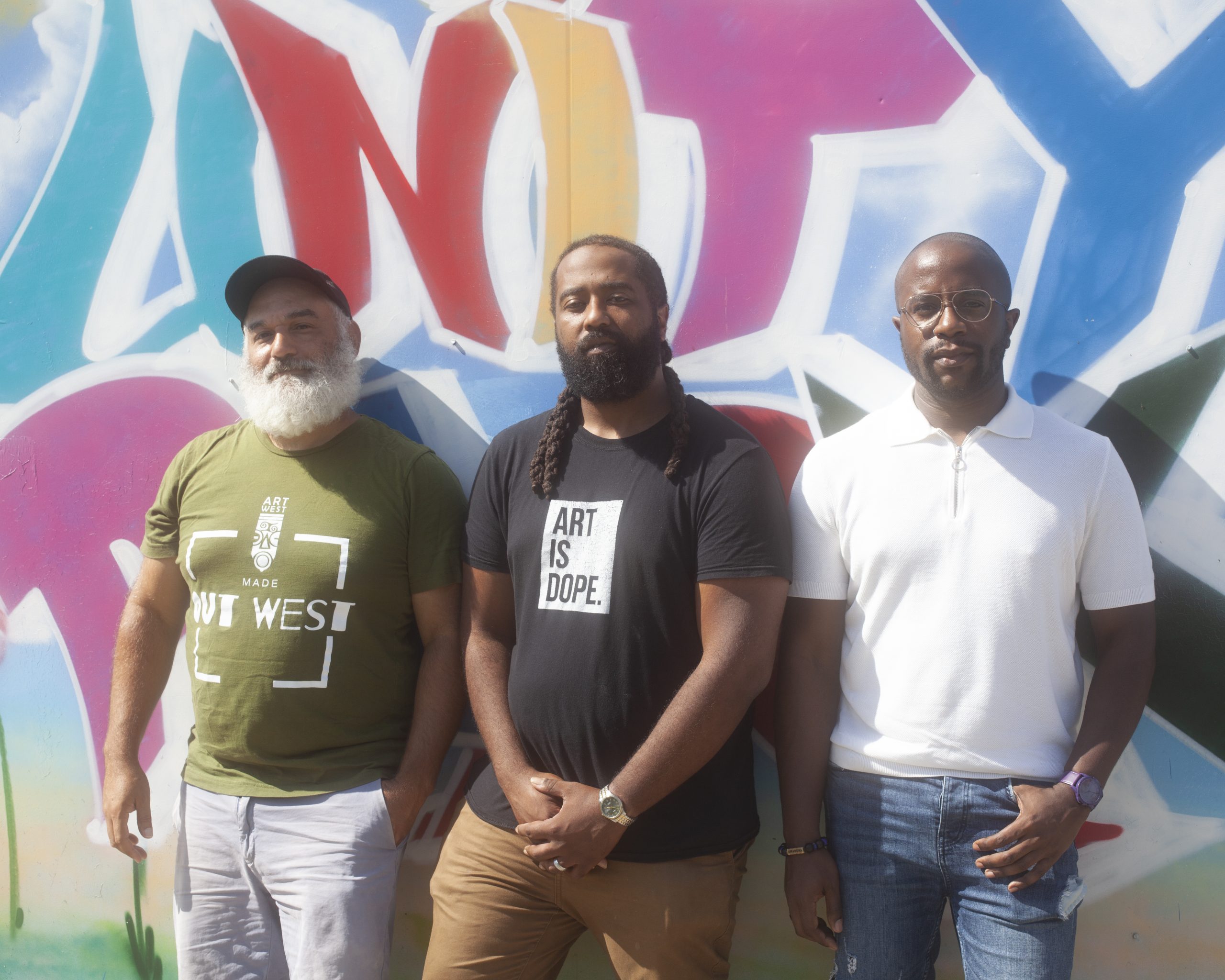 Image: Jonathan Kelley, Jay Simon, and Steven D. Booth stand side-by-side in front of a colorful mural with the word "Unity" painted just above their heads. They're each looking directly into the camera as the sun shines bright and high over them. Photo by Samantha Friend Cabrera.