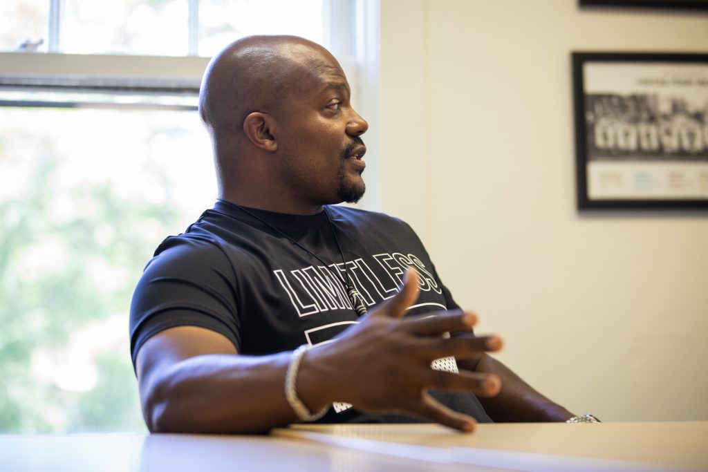 Image: Larry Brent Jr. seated at a table. Behind them is a window and to the right is a framed photograph on the wall. Larry looks off to the right and the fingers on his right hand are spread as he talks. Photograph by Kristie Kahns.