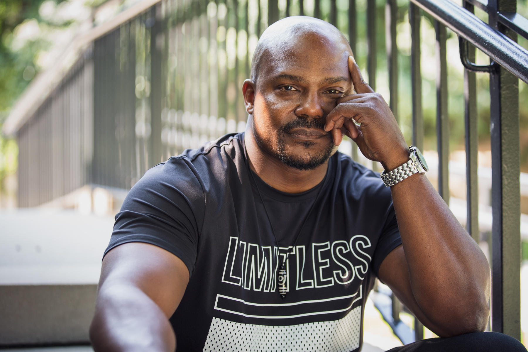 Featured image: Larry Brent Jr. on an outdoor concrete staircase next to metal railing. He wears a black t-shirt with the word "LIMITLESS" on it. His left hand is pressed up to his face, with his index finger on his temple. Photograph by Kristie Kahns.