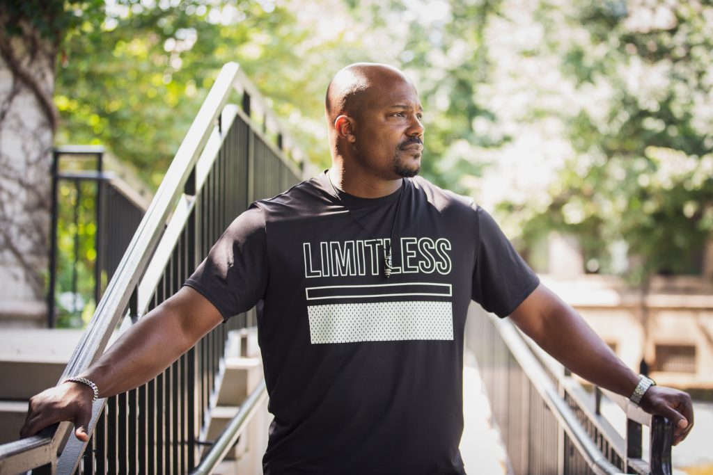 Image: Larry Brent Jr. stands on a concrete ramp his arms stretched to both sides, holding handrails. He looks off to the right and wears a black t-shirt with white text that says "LIMITLESS." In the background is bright greenery. Photograph by Kristie Kahns.