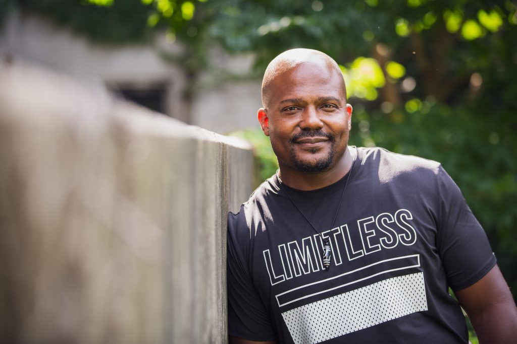 Image: Larry Brent Jr. leaning up against a concrete wall, about five inches shorter than him. Behind him, greenery. He wears a pendant and a black t-shirt with white text saying "LIMITLESS" on it. Photograph by Kristie Kahns.