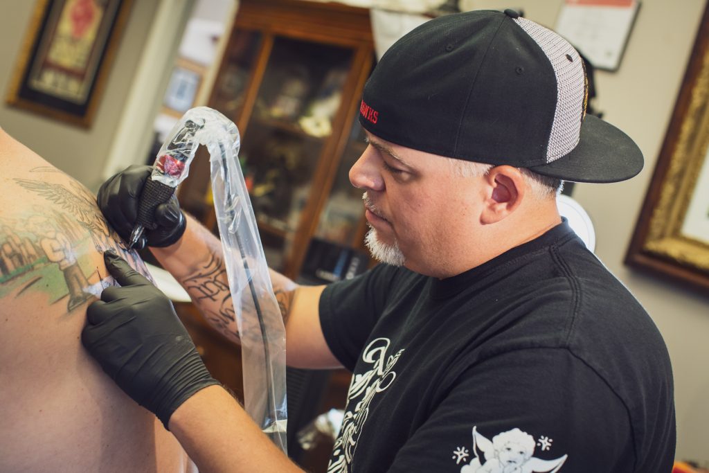 Image: Matt Sopron (right) works on a tattoo for a client at Dark Water Studio in Chicago, where he is on a tattoo apprenticeship. He wears black gloves and steadies his hands while creating an image on the client's back where we can partly see a scene of a young man at the Chicago lakefront on the left side, and on the right side is a winged figure holding scales of justice. Photograph by Kristie Kahns.