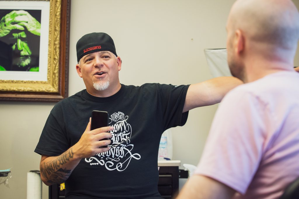 Image: Matt Sopron (left) talks with writer Michael Fischer at Dark Water Studio in Chicago. He is surrounded by tattooing equipment in the shop, and the walls have framed images of figures on them including a green-hued image of a distraught-looking man with his hands on his head. Photograph by Kristie Kahns.