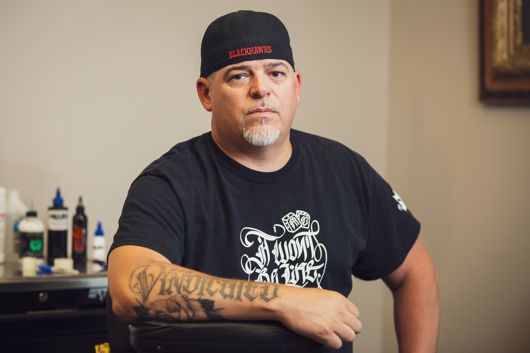 Image: A portrait of Matt Sopron at Dark Water Studio in Chicago, where he is on a tattoo apprenticeship. He wears a black t-shirt and backwards hat. His right arm rests on the arm of the chair, and partially visible is a large tattoo that says "Vindicated" on his arm. Photograph by Kristie Kahns.