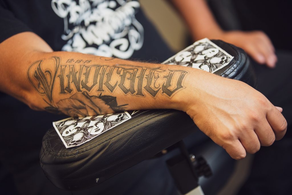 Image: A close-up detail of a tattoo on Matt Sopron's right arm, which says "Vindicated" and includes the date 12/18/18 when he was exonerated. His arm is on a black leather arm rest and is surrounded by black and white drawings of skulls and long-haired figures. Photograph by Kristie Kahns.