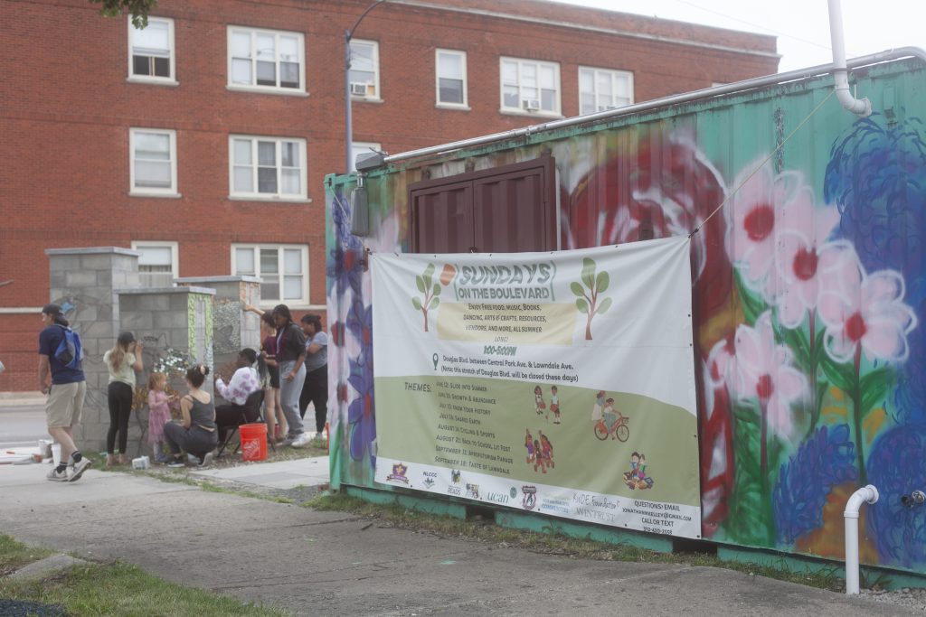 Image: People can be seen in the distance working together on an outdoor mosaic installation. Nearby is a shipping container that has a mural painted on the outside. A banner hangs on the container, with the words Sundays on the Boulevard in green letters across the top. Photo by Samantha Friend Cabrera.