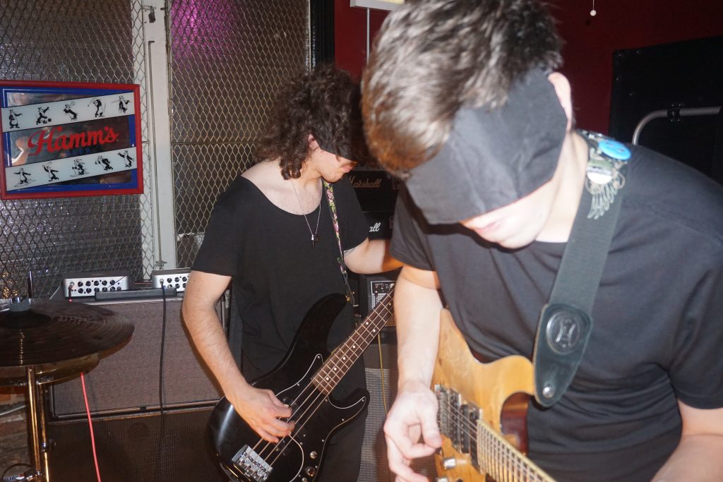 Image: At Reed's Local, two blindfolded guitarists are playing. One is blurry in the foreground, the other is in-focus in the back. Behind them on a counter is some sound equipment. Hanging from the wall is an old sign for Hamm's beer. Photograph by Hector Frias.