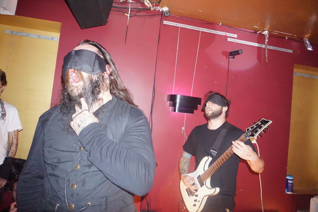 Image: In a red and golden room, two bearded men are blindfolded. The one on the right plays a white electric guitar. The one on the left opens his mouth wide and has a hand in his beard. Photograph by Hector Frias.