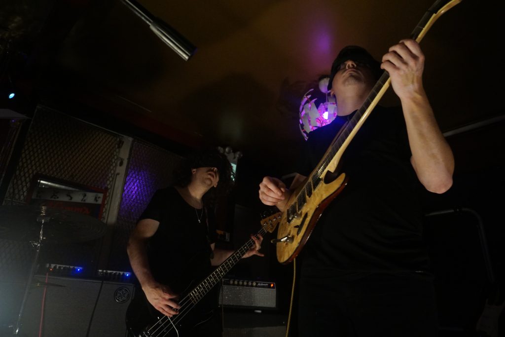 Image: In a darkened room, a low-angle photo of two guitarists that are next to each other. Behind them are amplifiers with glowing lights and a dim exit sign. On the ceiling is a white lamp with a purple bulb in it. Photograph by Hector Frias.