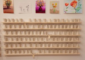 Image: Above six wooden shelves of ceramic cups styled like styrofoam cups, are five paintings of flowers by Abdualmalik Abud, Djamel Ameziane, Ghaleb al-Bihani, and Assad “Haroon” Gul. Photo by Zoey Dalbert. Courtesy of the DePaul Art Museum.