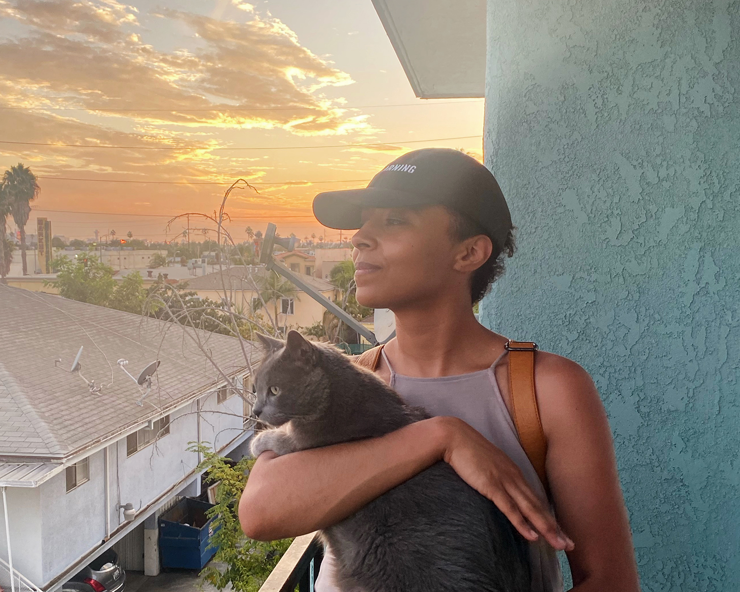 Image: A portrait of Kiki holding their cat Momi in their arms, shown from the waist up. Kiki and Momi are looking away from the camera and gazing out toward the cityscape. They are observing the view from a balcony at home. The sun sets behind them casting warm colors of orange, pink and purples into the sky among the clouds. Photo by Matthew Austin.