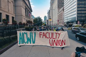 Image: On the sidewalk in front of the Art Institute of Chicago, a line of protestors face the camera holding a white cloth banner that says in blue and red text "AICWU Faculty Union." The sky is clear behind them. Photo by Apollo Nava.