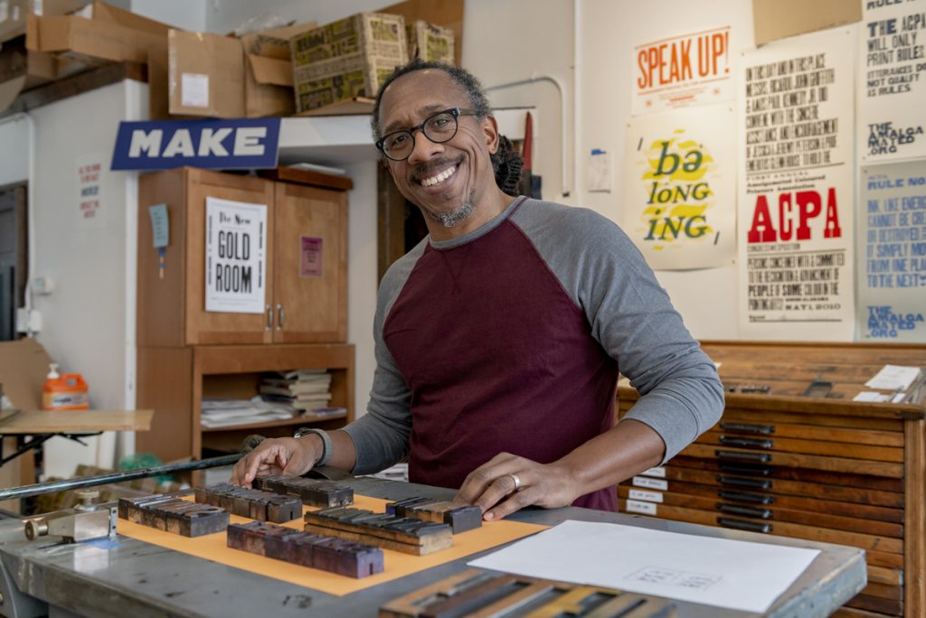 Right Image: Artist Ben Blount smiles at the camera while standing by a letterpress in his studio. Photo by Ryan Edmund Thiel.